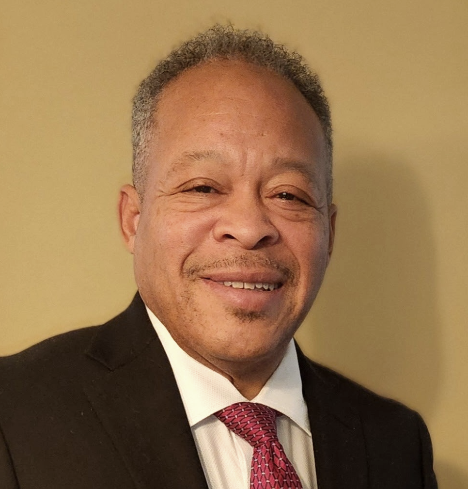 Lee Coleman, smiling and in a suit, with a tie, standing against a plain background.